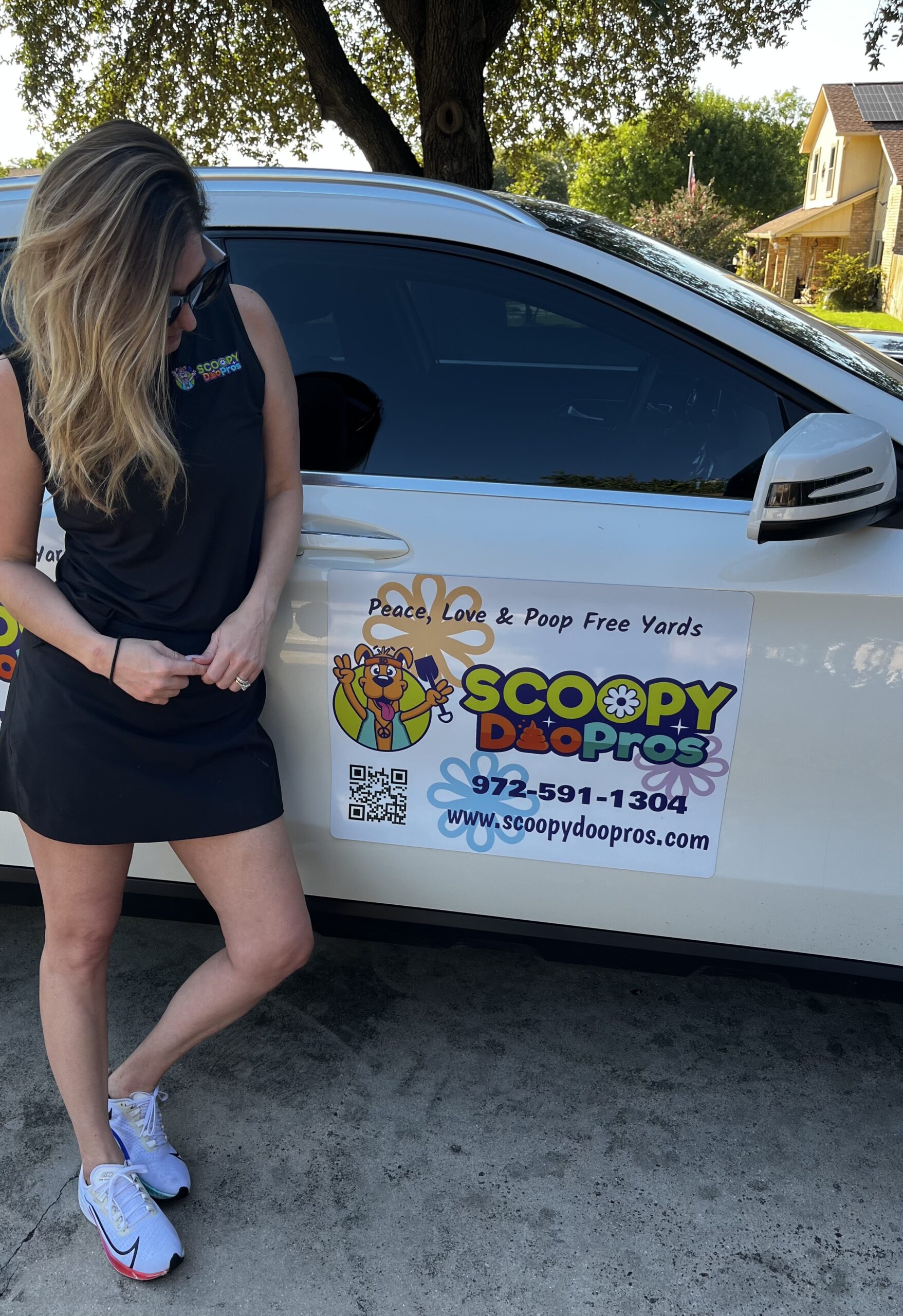 A woman wearing a black dress and sunglasses stands next to a white vehicle with a Scoopy Doo Pros decal on the side. The decal displays the company's logo, contact information, and tagline, "Peace, Love & Poop Free Yards." The logo features a cartoon dog with a colorful, groovy design.